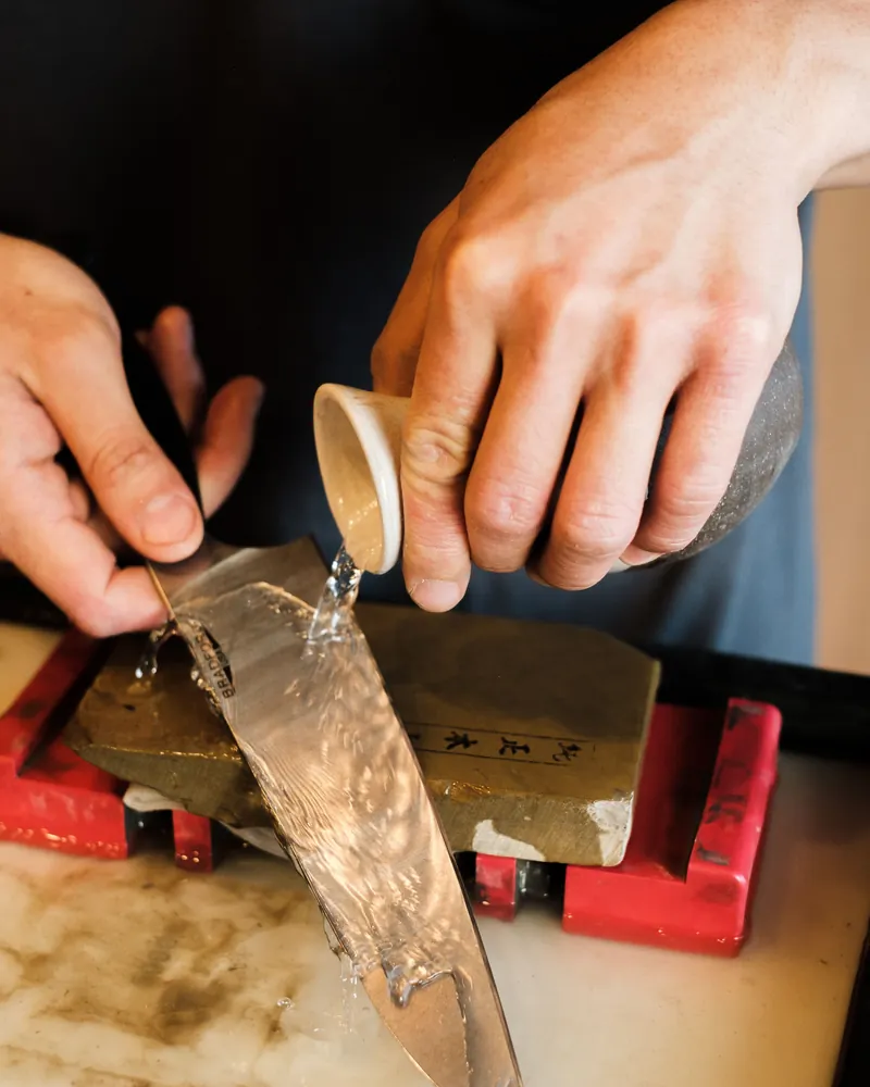 Manos afilando la hoja de un cuchillo sobre una piedra de agua mientras se vierte agua sobre ella